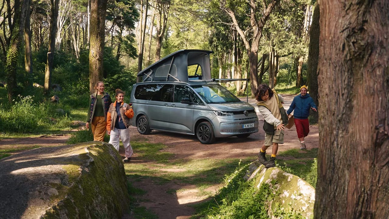 Un camper Volkswagen California in un bosco con tenda alzata e persone
